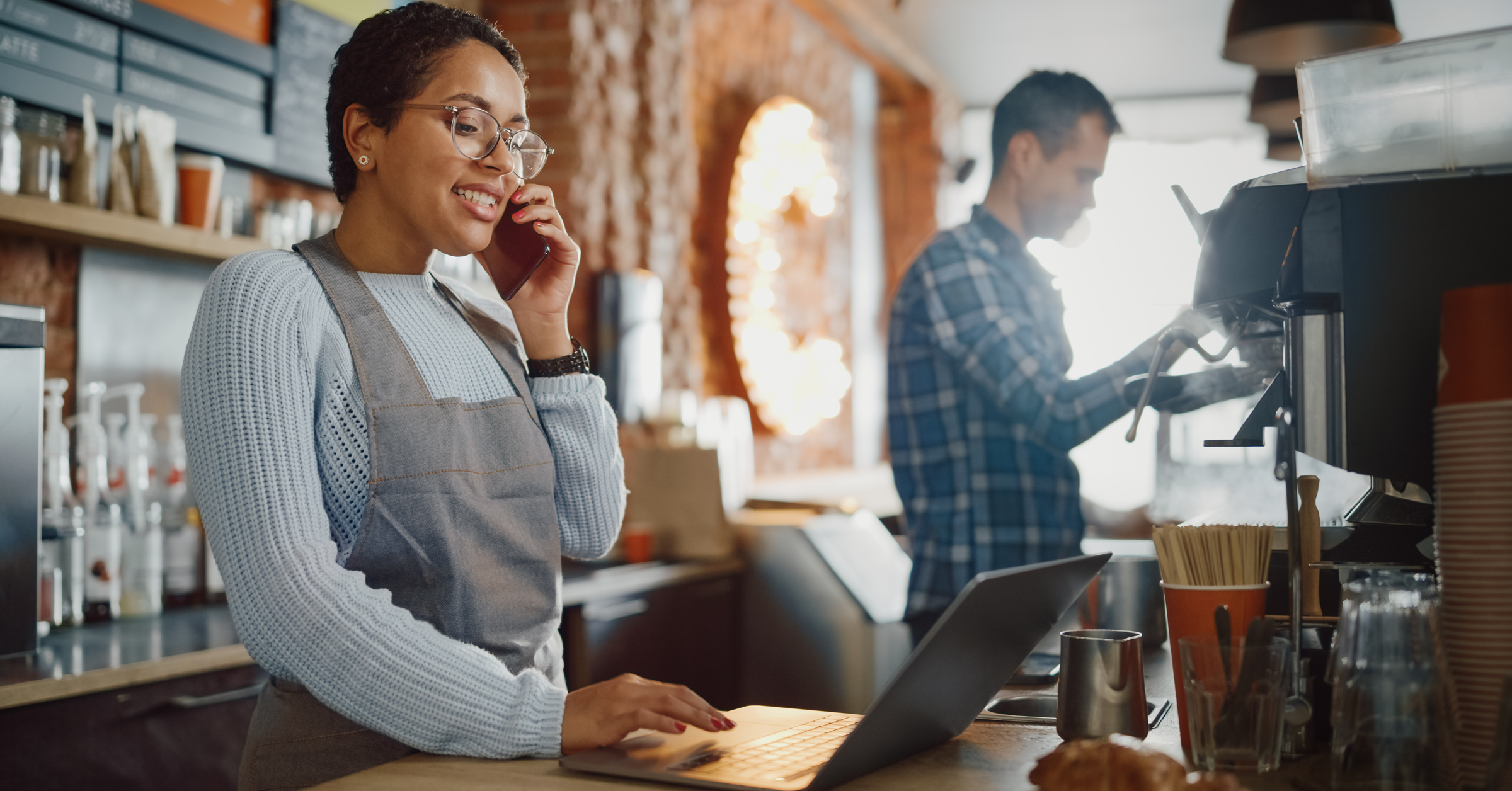 Cafe owner standing with laptop on bench and talking on mobile